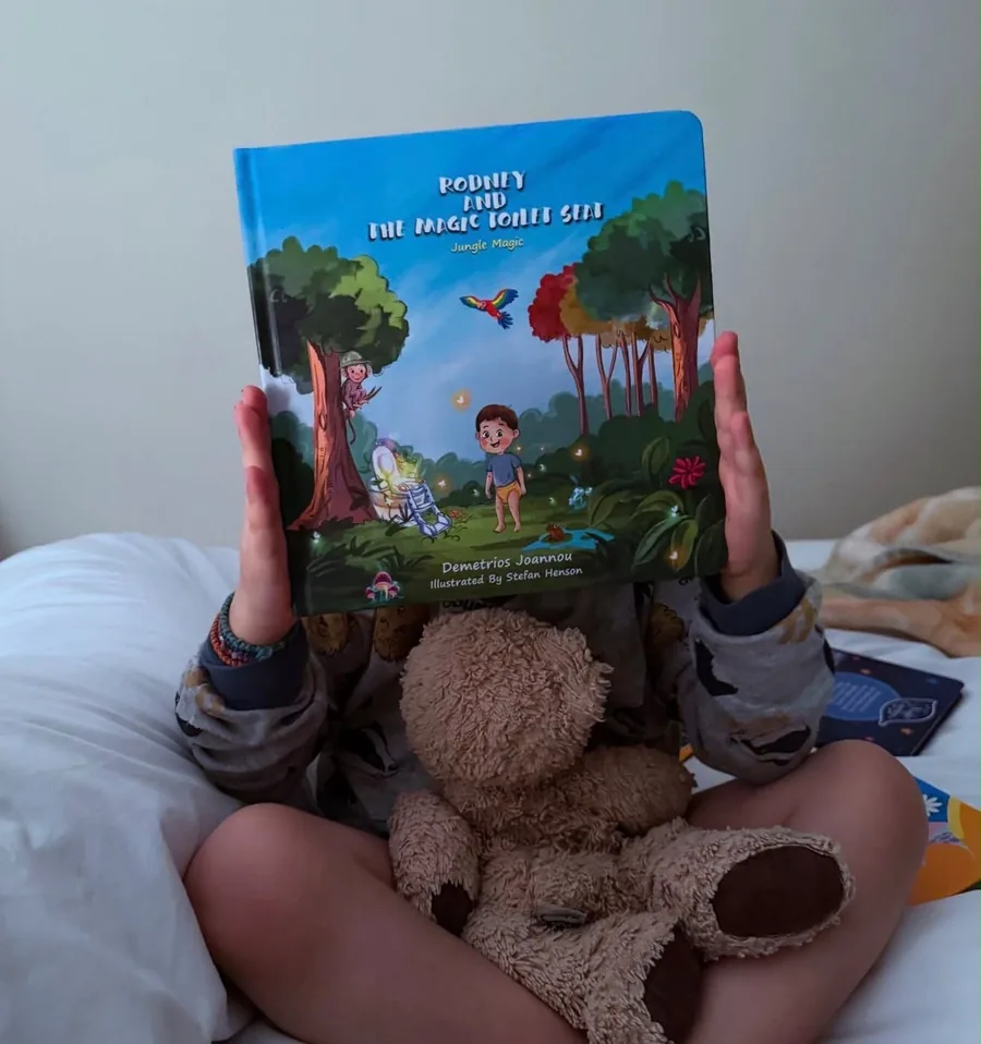 Niko sitting on his bed holding up Rodney and the Magic Toilet Seat, teddy on his lap