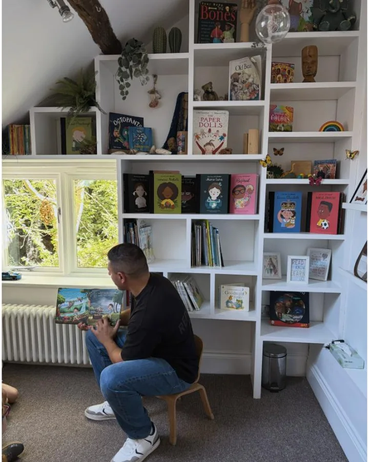 Demetrios reading the book aloud to a nursery class beside a full bookshelf